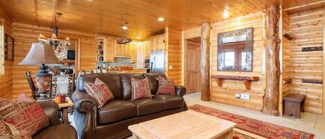 A cozy log cabin interior with a leather sofa, wooden coffee table, and a kitchen area.