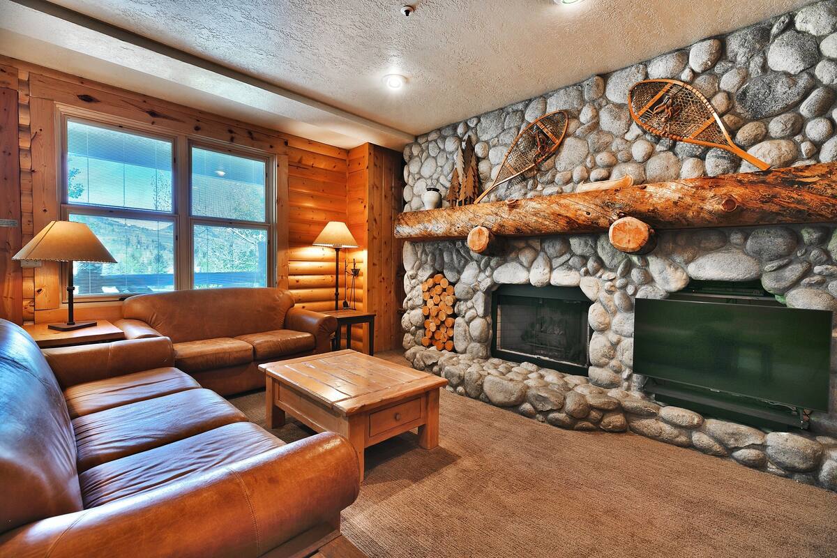 A log cabin interior with a stone fireplace, leather sofas, and a television.