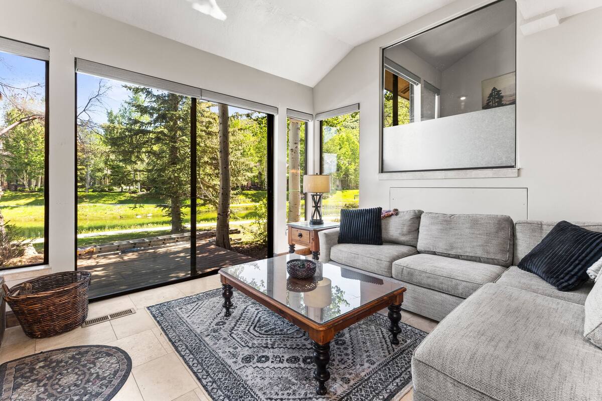 A modern living room with a large glass door leading to a garden, a grey sofa, a wooden coffee table, and a patterned rug.