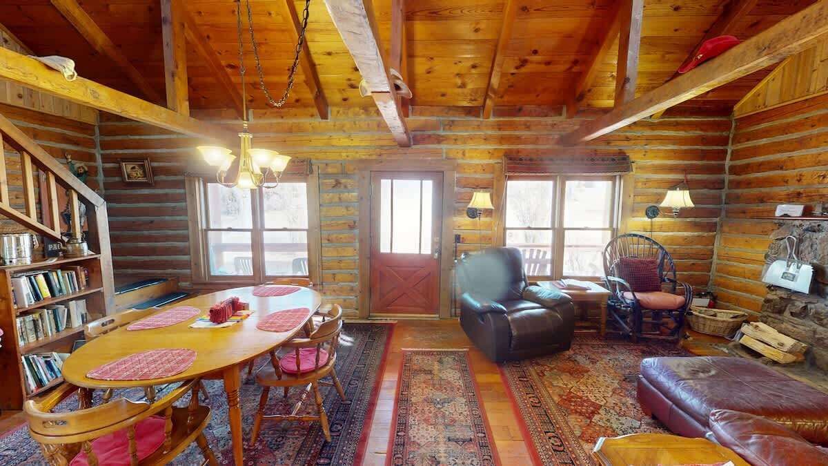 A log cabin interior with a dining table, chairs, a leather sofa, a rocking chair, and a bookshelf.