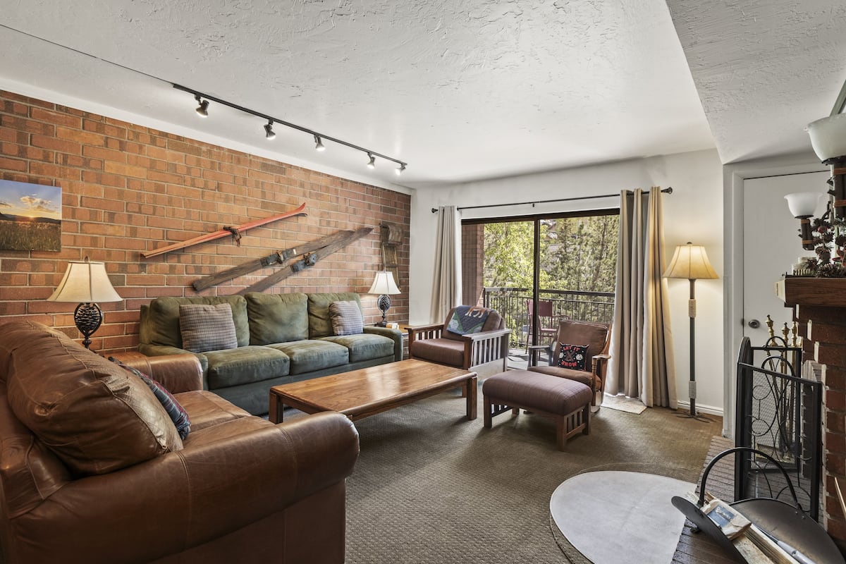 Een gezellige woonkamer met een bakstenen muur, een leren bank, een houten salontafel en een balkon met uitzicht op bomen.