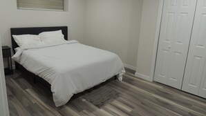 A bedroom with a bed, a nightstand, a window with blinds, a door, and a wooden floor. - Cozy 2-Bedroom basement Suite with Private entrance South Winnipeg. (Winnipeg)