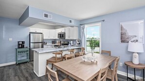 A modern kitchen with white cabinets, stainless steel appliances, and a marble countertop. There is a dining area with a wooden table and chairs, and a view of the outdoors through a large window.