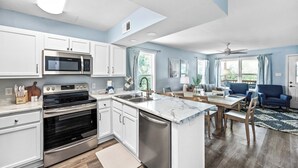 A modern kitchen with white cabinets, a built-in microwave, and a large island. There is a dining area with a table and chairs, and a living room with a blue-striped sofa.