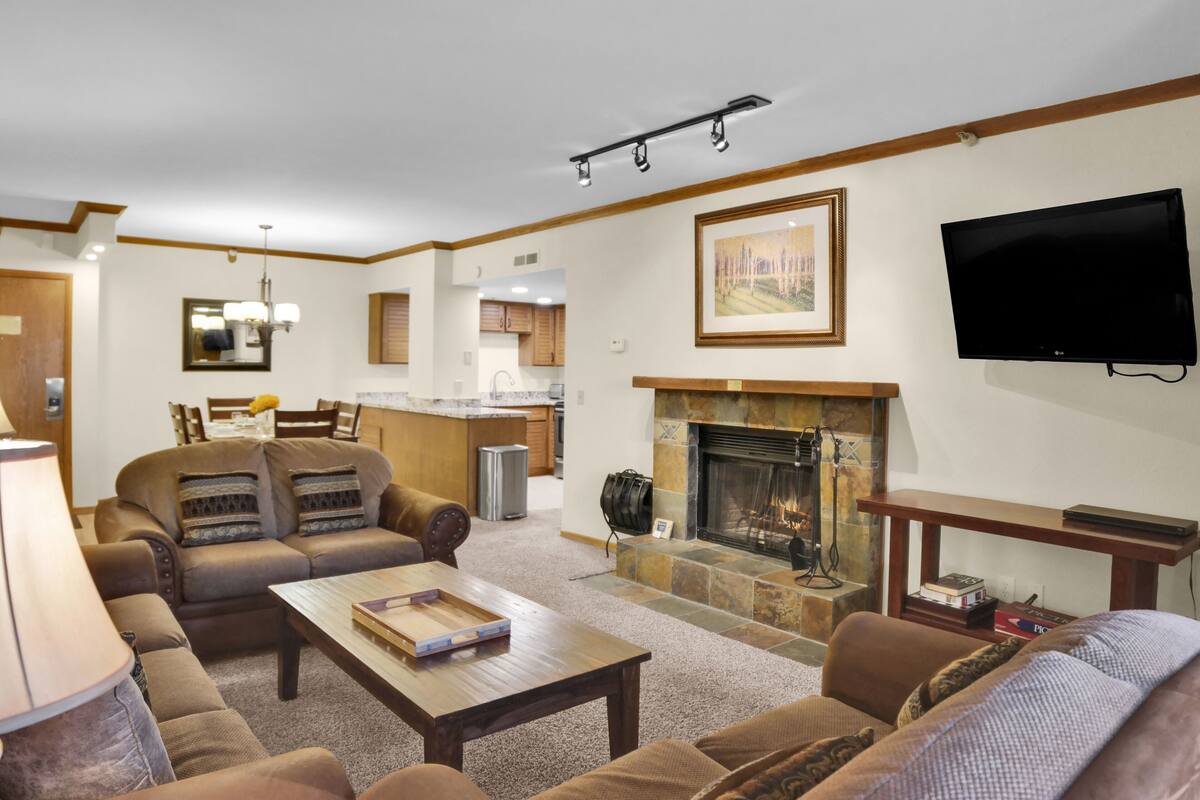 A spacious living room with a fireplace, a television mounted on the wall, and a kitchen in the background.
