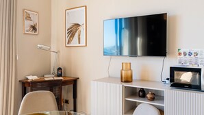 Une chambre d’hôtel moderne dotée d’un téléviseur à écran plat fixé au mur, d’un bureau en bois avec une lampe, d’une table en verre avec un bol et d’une armoire blanche agrémentée d’objets décoratifs.