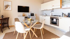 Une cuisine moderne avec des meubles blancs, un coin repas doté d’une table et de chaises en verre, un téléviseur à écran plat et un four à micro-ondes.