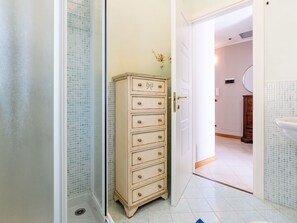 Un baño con piso de baldosas blancas, un aparador beige con varios cajones, una zona de ducha con puerta de vidrio y un lavamanos blanco con espejo.