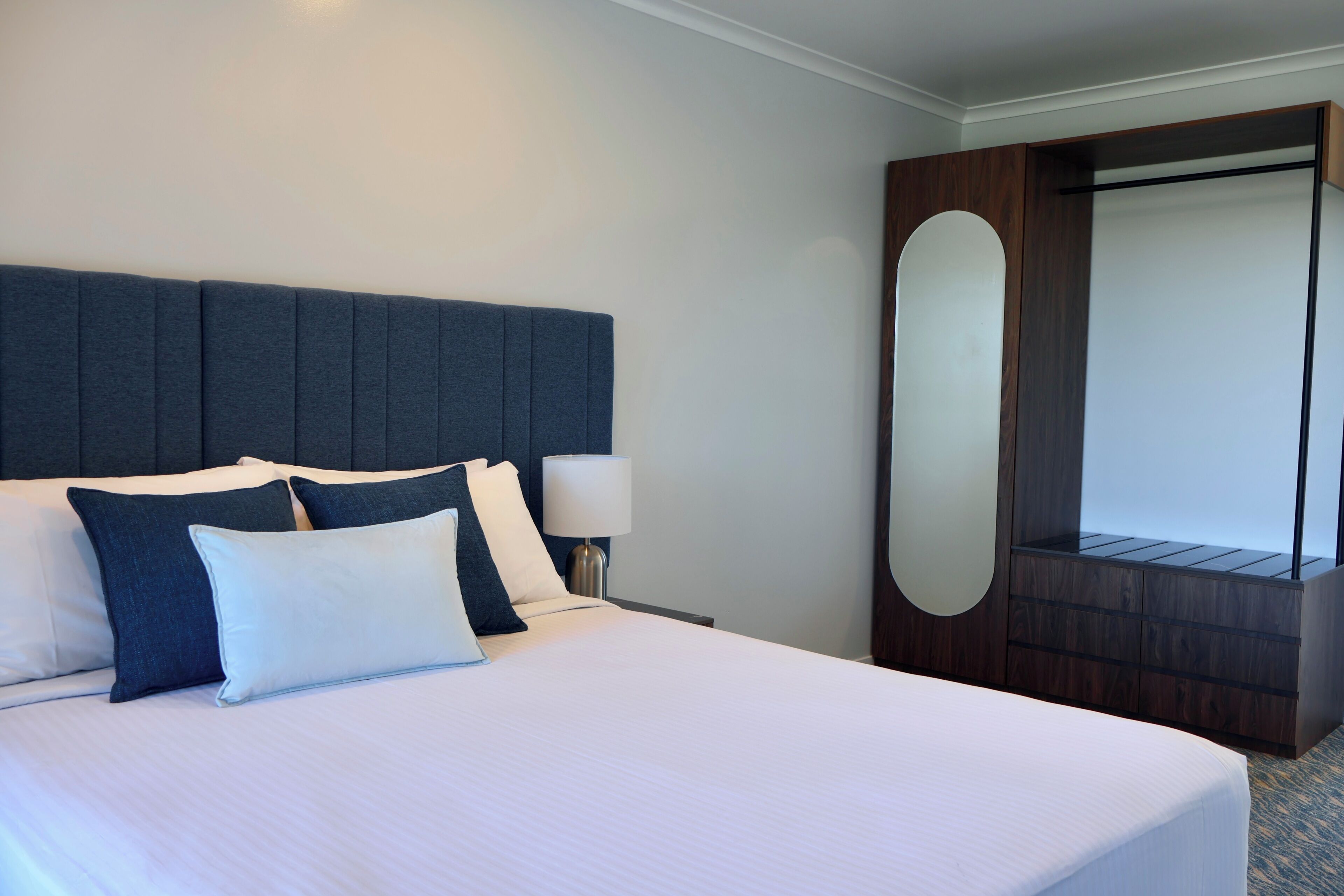 Una habitación de hotel con una cama grande, cabecera, mesita de noche y un espejo.