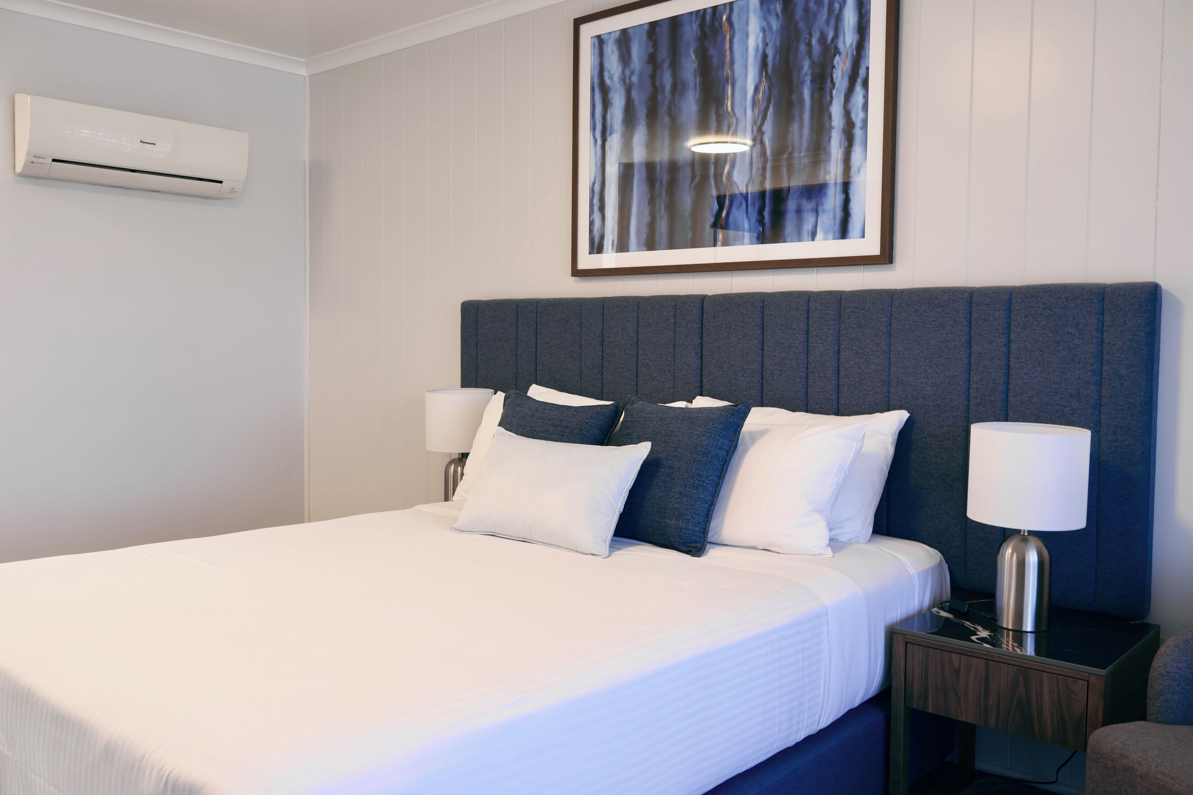 Habitación de hotel con cama, lámparas de noche, mesita de noche y un cuadro en la pared.