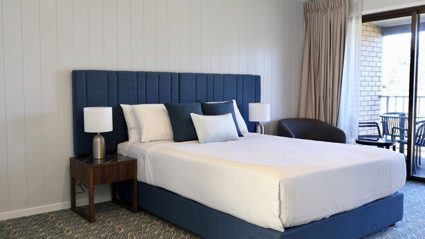 A hotel room with a large bed, two bedside tables with lamps, a view of the outside balcony, and a patterned carpet. - Victory Hotel Motel Gympie  (Gympie)