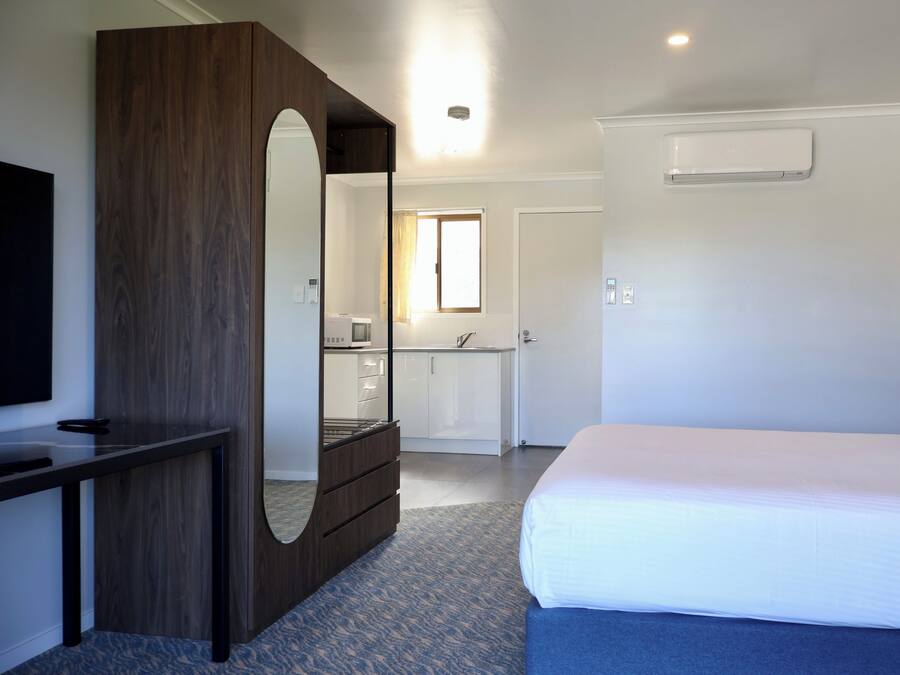 Un dormitorio con cama, cómoda, espejo, televisor y baño visible a través de una puerta abierta.