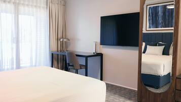 Une chambre d’hôtel avec un grand lit, un bureau, une chaise, un téléviseur à écran plat, un miroir et une vue sur l’extérieur à travers des rideaux transparents.