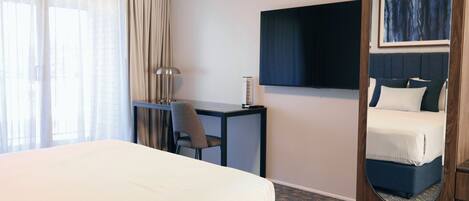 Een hotelkamer met een groot bed, een bureau, een stoel, een flatscreen televisie, een spiegel en uitzicht buiten door dunne gordijnen.
