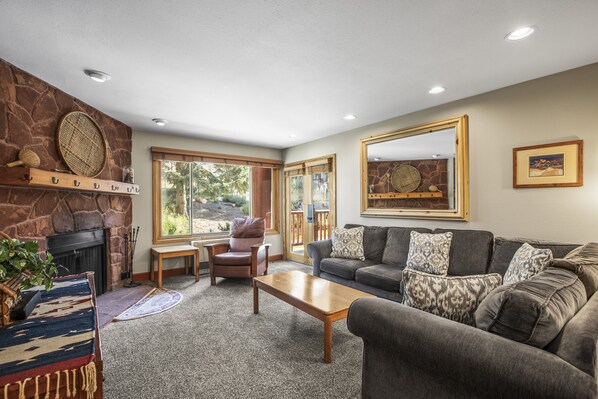 A cozy living room with a stone fireplace, a large window, a gray sofa, and a wooden coffee table.
