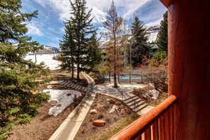 A view from a wooden balcony overlooking a landscaped area with a pool, trees, and a building in the background.