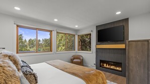 Een moderne slaapkamer met een groot bed, een open haard, een televisie en grote ramen met uitzicht op bomen.