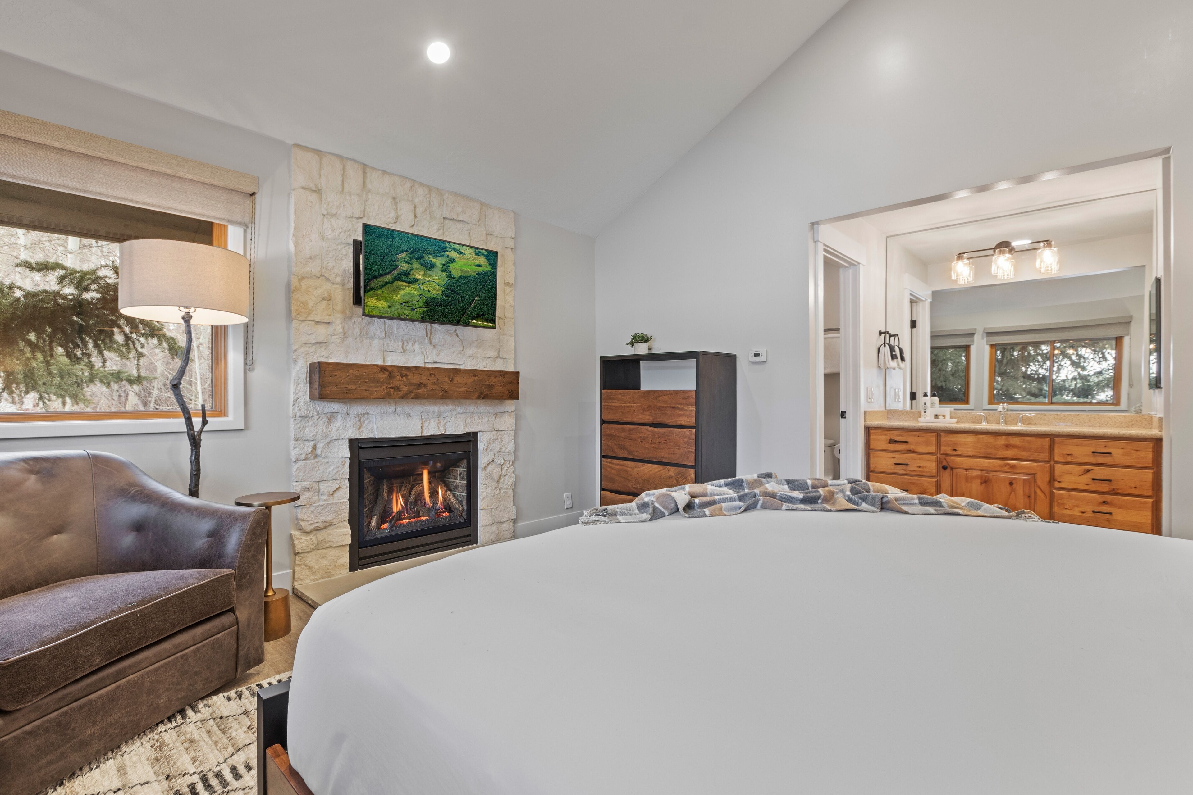 A bedroom with a fireplace, a large bed, a sofa, and a bathroom with a mirror and sink.