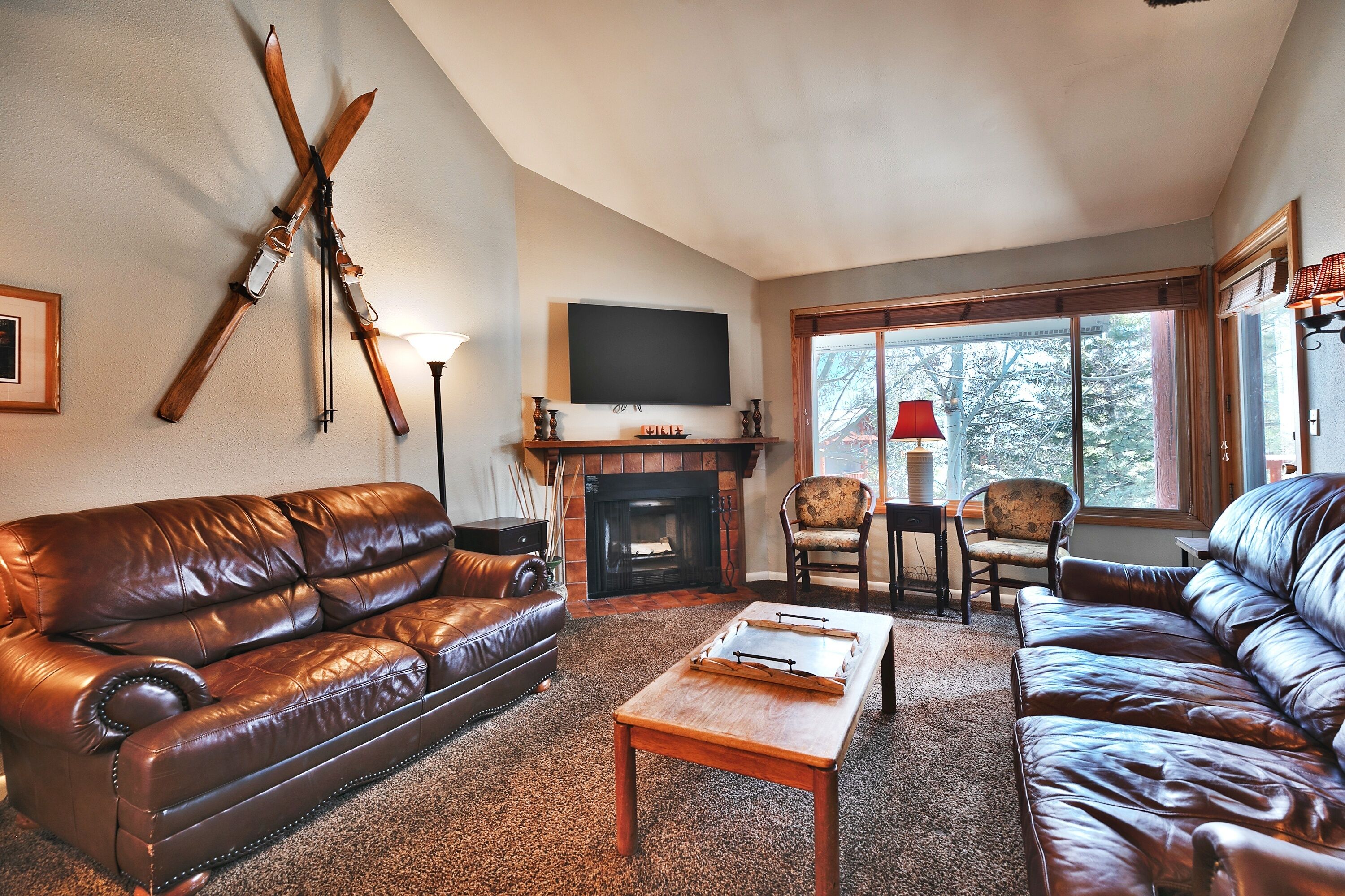 A cozy living room with leather sofas, a fireplace, a mounted TV, and a window with a view of trees.