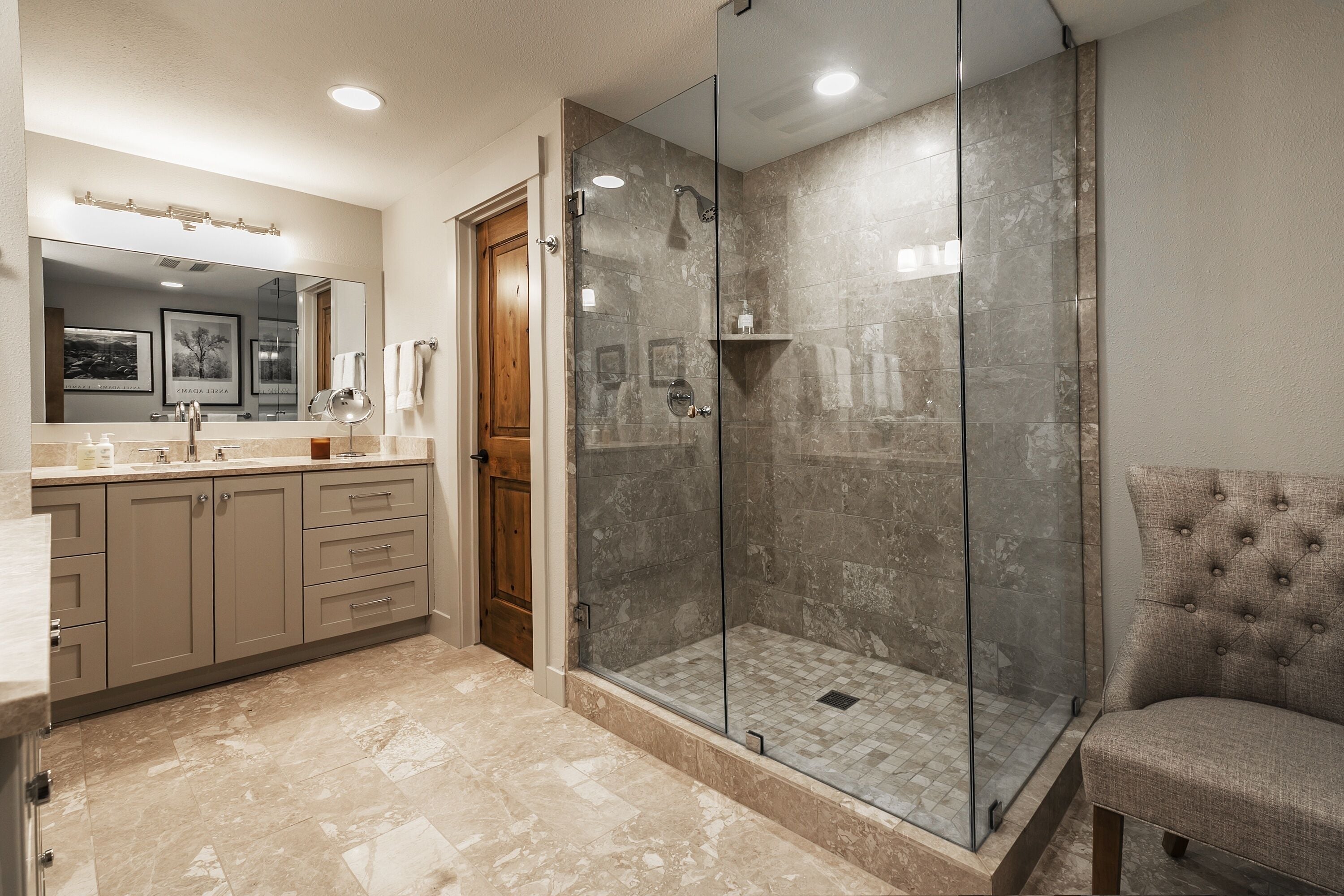 Un baño moderno con ducha de vidrio, tocador con espejo y sillón tapizado.