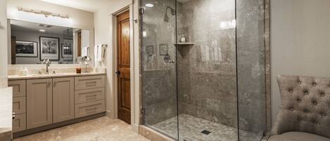 Un baño moderno con ducha de vidrio, tocador con espejo y sillón tapizado.