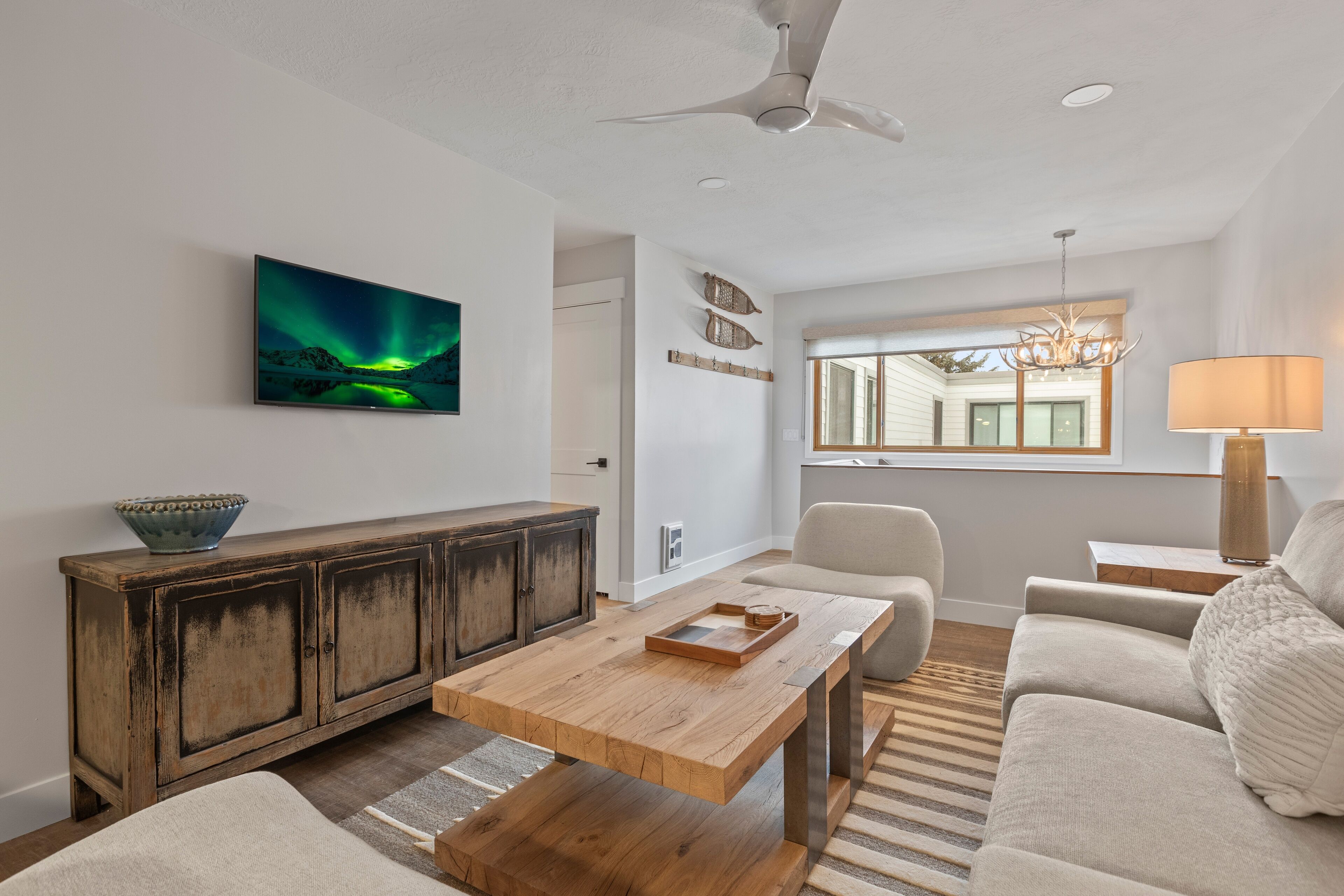 Un salon moderne avec une table basse en bois, un canapé, un meuble, un lustre et une fenêtre donnant sur une maison.