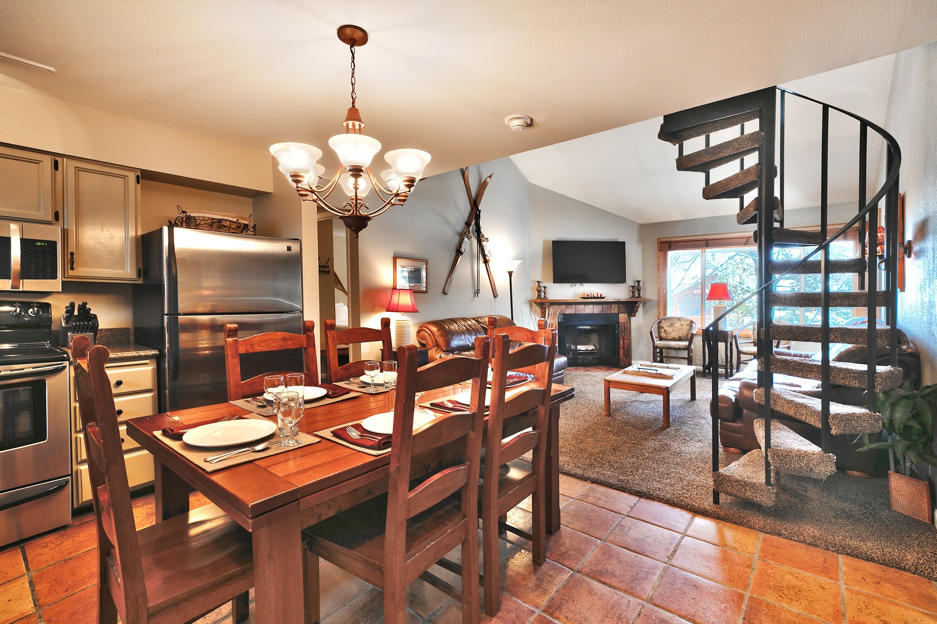 A dining area with a wooden table and chairs, a chandelier, and a kitchen with stainless steel appliances.