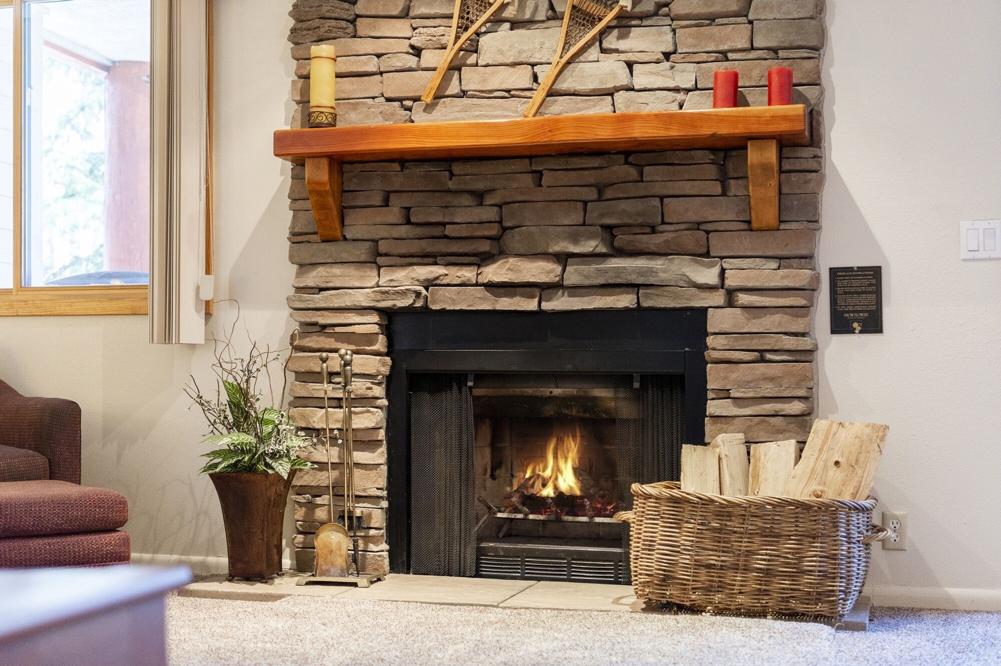 A stone fireplace with a wooden mantel shelf, a fire burning, and a basket of wood logs.