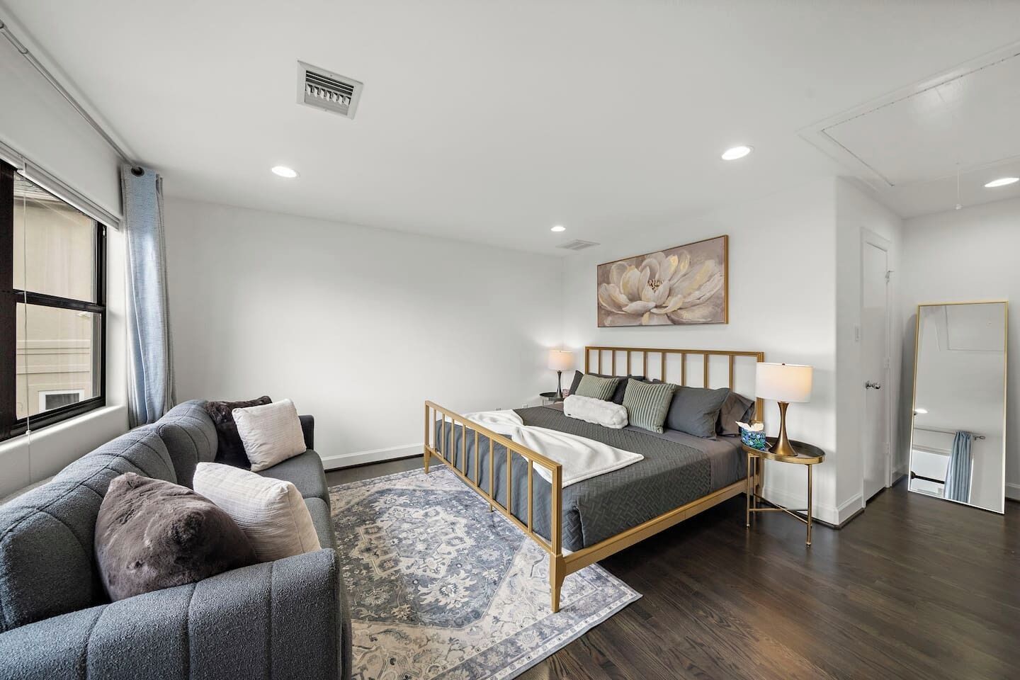 A modern bedroom with a bed, a sofa, a bedside table with lamps, a large mirror, and a window with curtains.