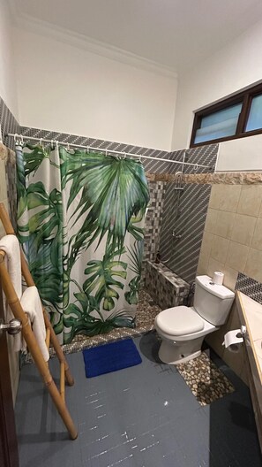 Une salle de bain avec un rideau de douche orné d’un motif de feuilles tropicales, des toilettes, un bidet et un petit tapis bleu sur le sol.