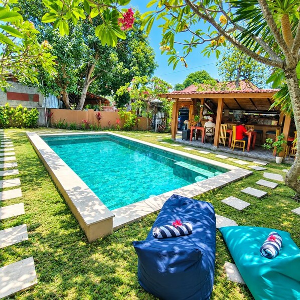 Une piscine dotée d’une terrasse carrelée, entourée de pelouse et de plantes, avec un espace salon couvert et un bar en arrière-plan.