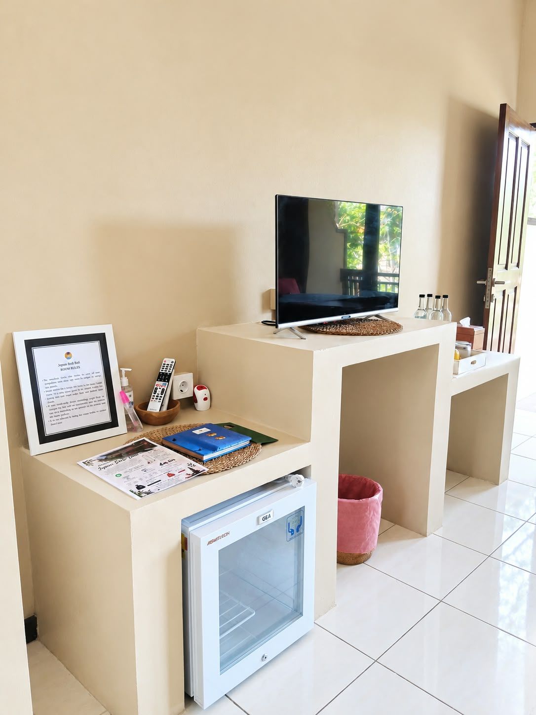 Zona de recepción de un hotel con televisión, cartel de bienvenida enmarcado, controles remotos, un electrodoméstico GE y un cesto de basura.