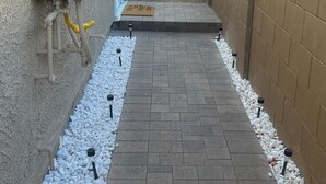 A narrow pathway with a brick floor, surrounded by white pebbles and a metal grate at the bottom.
