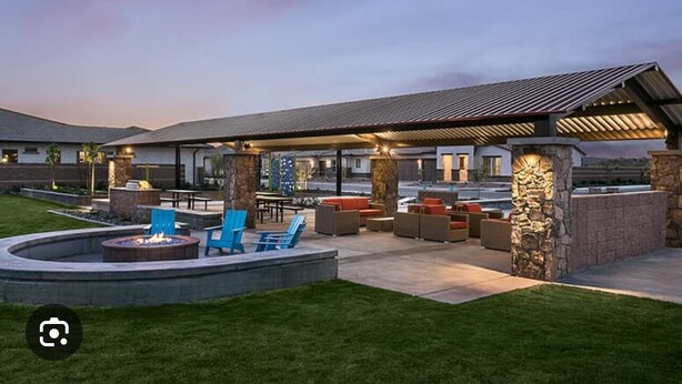 A covered outdoor patio with seating and a fire pit, surrounded by stone pillars and a well-maintained lawn.