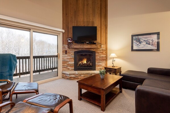 Cozy living room with a fireplace, comfortable seating, and a TV above, offering a view of the outdoors.