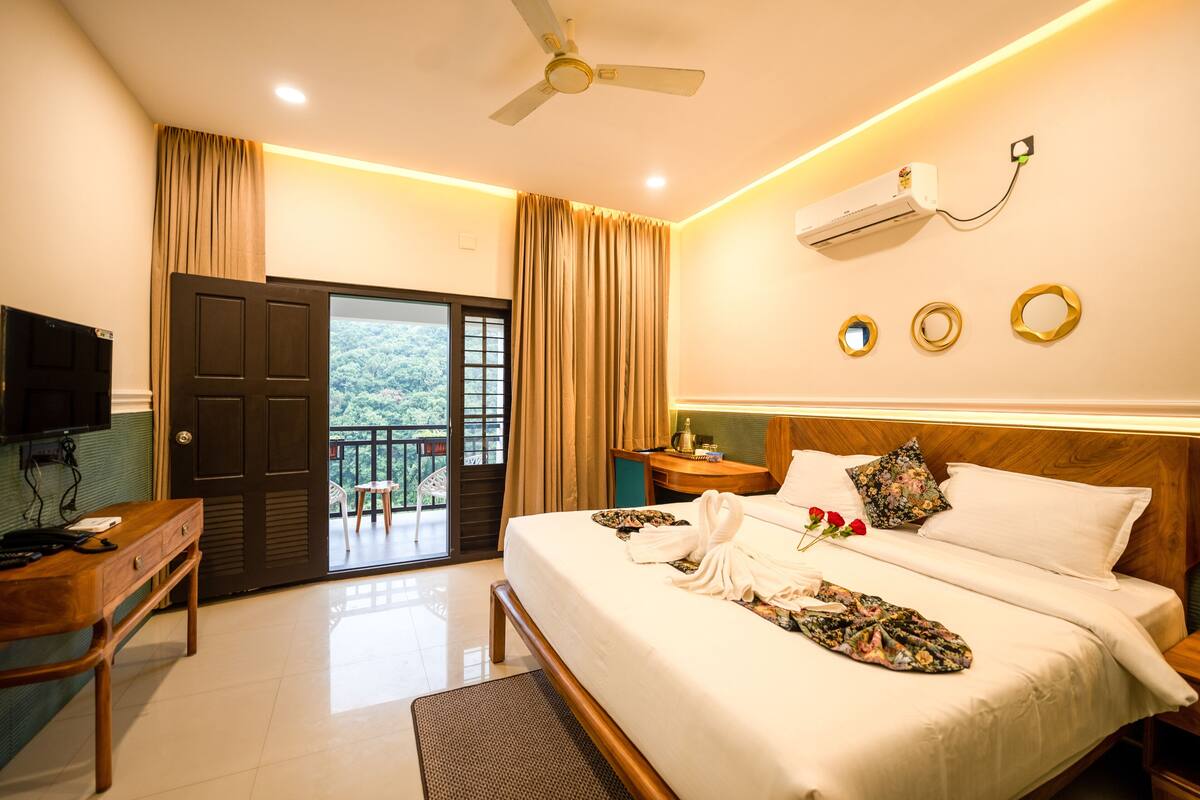 A hotel room with a bed, a desk, a television, an air conditioning unit, and a balcony with curtains.