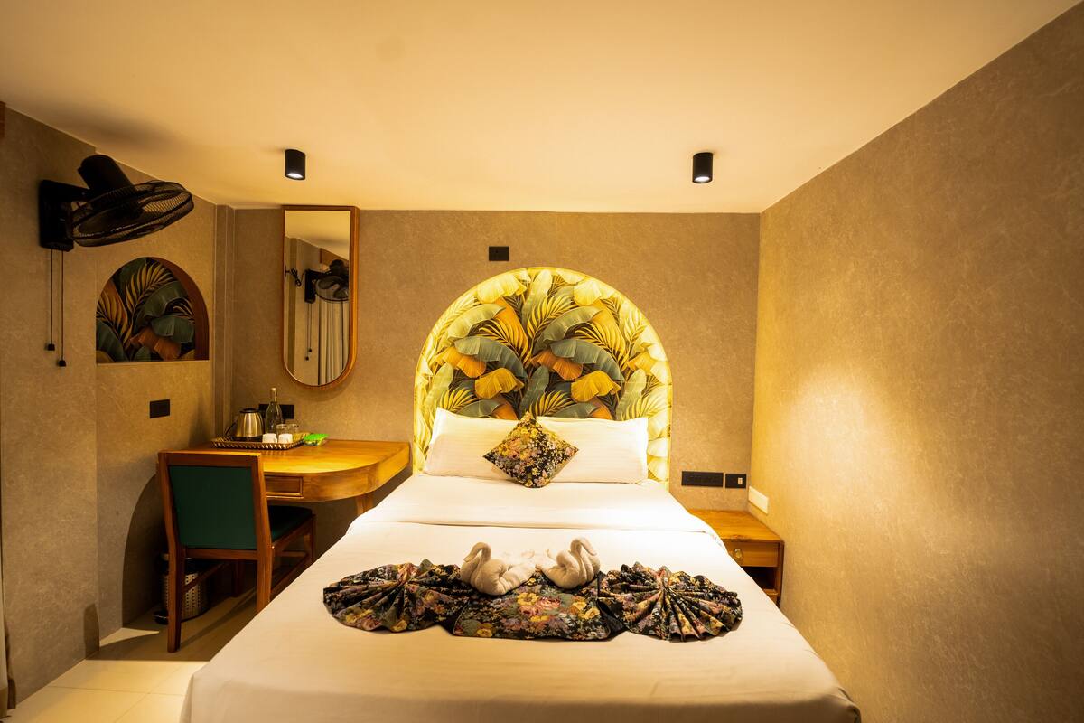 A hotel room with a bed, a desk, a chair, a mirror, and a wall-mounted fan.