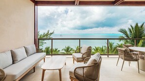 A balcony with a sofa, chairs, and a table, overlooking a turquoise sea and palm trees.