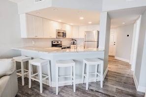 A modern kitchen with white cabinets, a central island with bar stools, stainless steel appliances, and a wooden floor. - Seasalt Shores - Oceanfront Condo (Ponce Inlet)