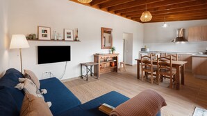 Ein modernes Wohnzimmer mit einer Couch, einem Fernseher, einem Essbereich mit Holzstühlen und einer Küche mit weißen Schränken.