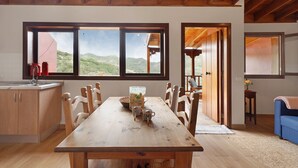Ein Holzespeisesaal mit Stühlen, eine Küchenzeile mit Schränken und große Fenster mit Blick ins Freie.