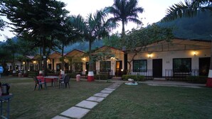 A courtyard with outdoor seating, palm trees, and a pathway leading to a building with multiple doors. - Antaram Resort (Rishikesh)