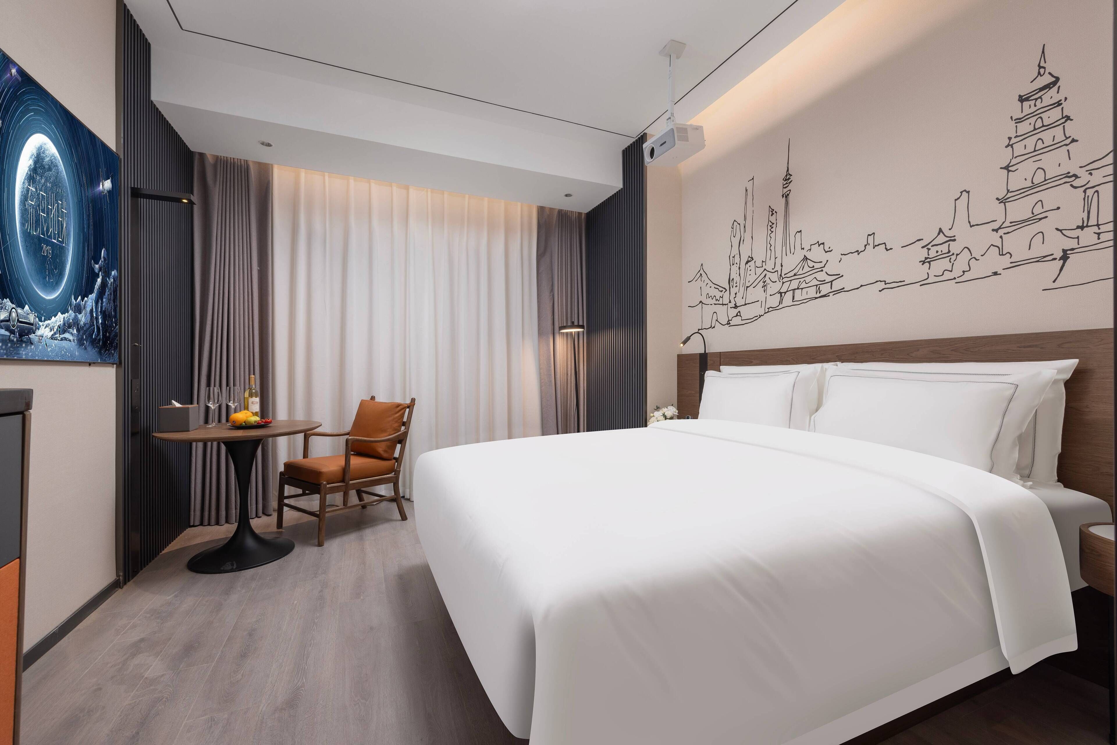 A hotel room with a large bed, a small table with a chair, a projector screen, and a wall mural.