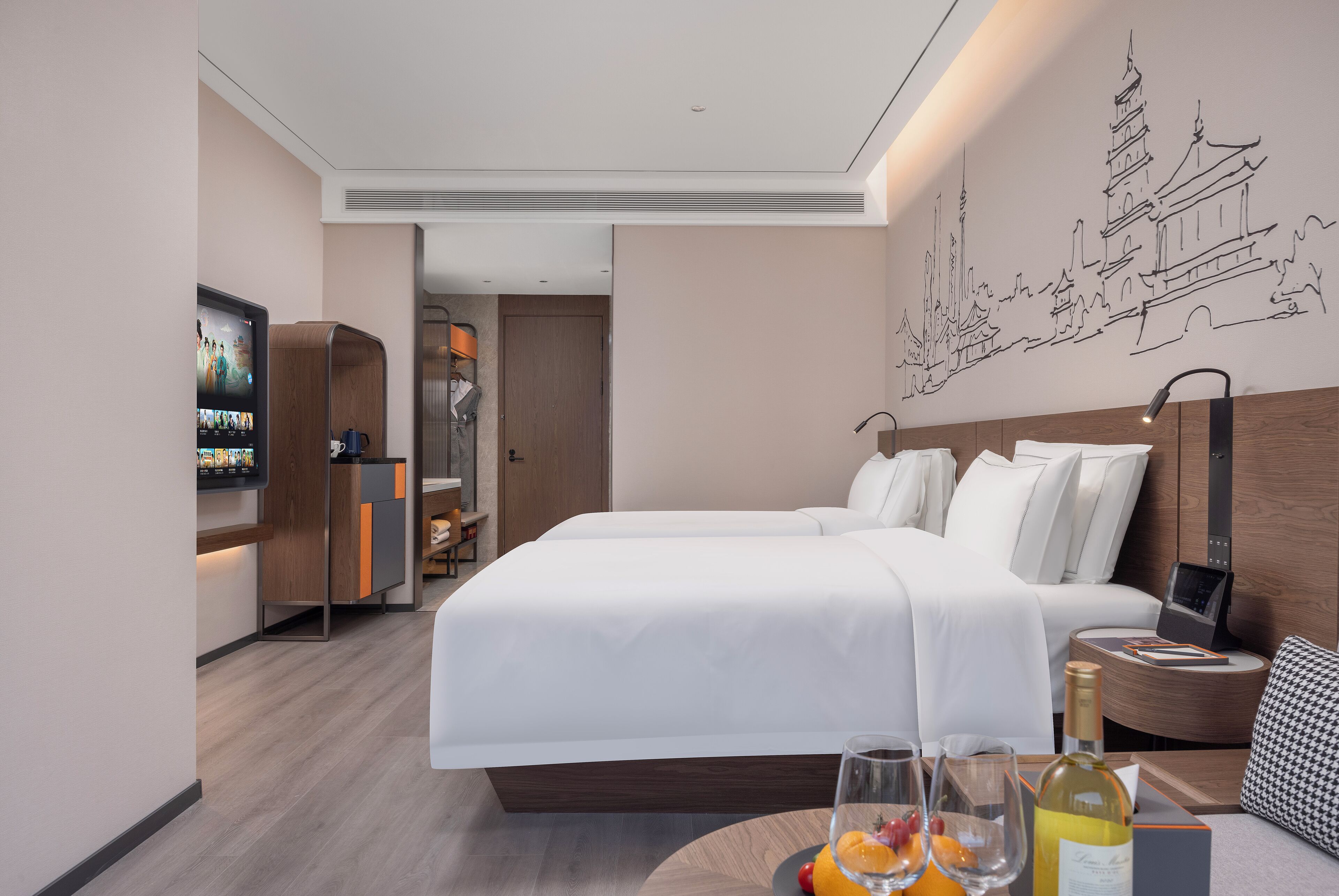 Una habitación de hotel moderna con una cama grande, una mesita de noche con una botella y vasos, un televisor y una obra de arte en la pared que representa un paisaje urbano.