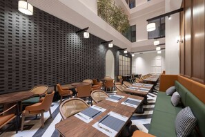 Uma área de refeições de hotel moderna, com mesas comunitárias grandes, assentos acolchoados e um piso estampado.