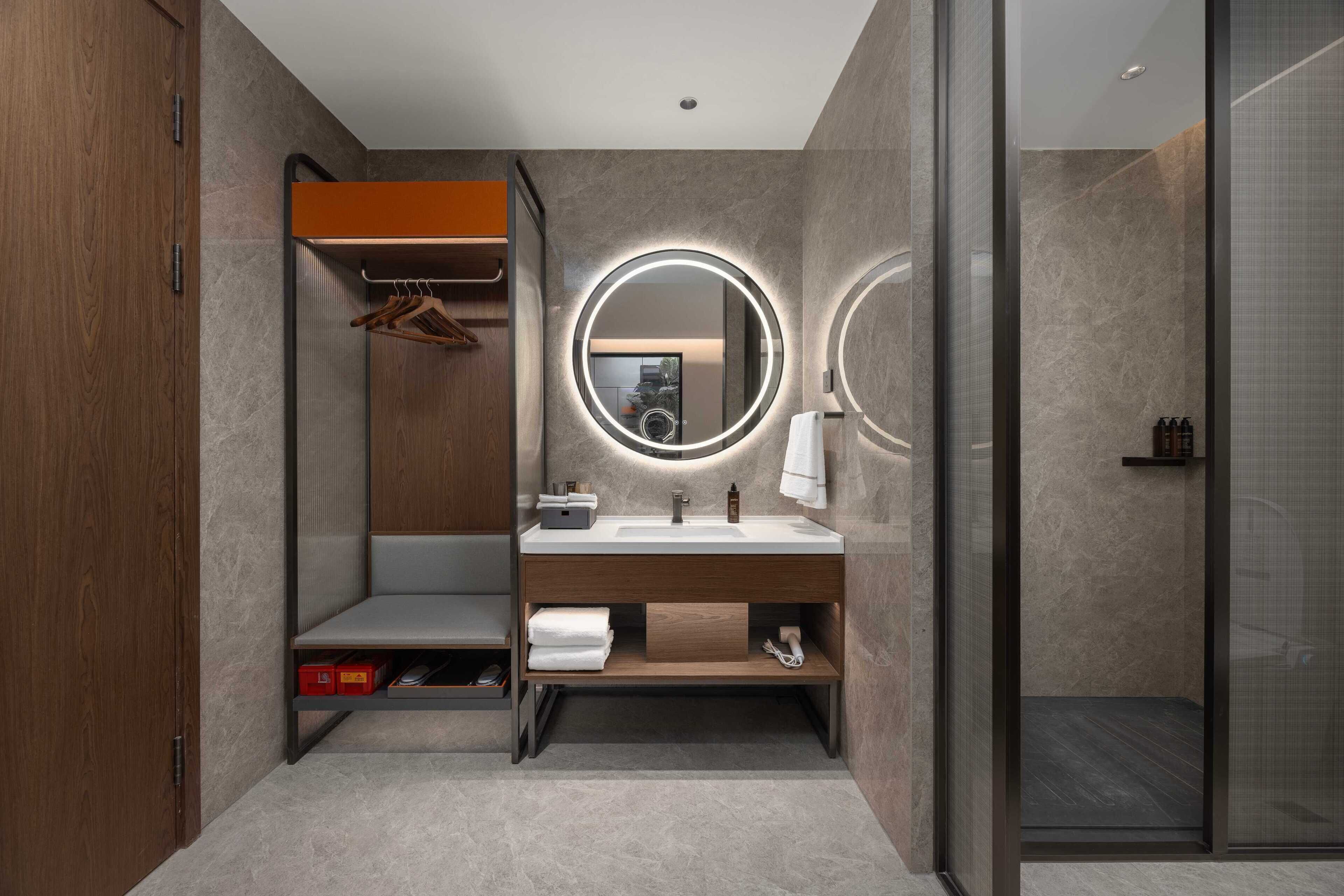 A modern bathroom with a wooden vanity, a round mirror with underglow lighting, a glass shower enclosure, and a built-in bench.