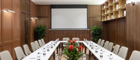 Un salón de conferencias con una larga mesa, sillas, una pantalla de proyector y una estantería.