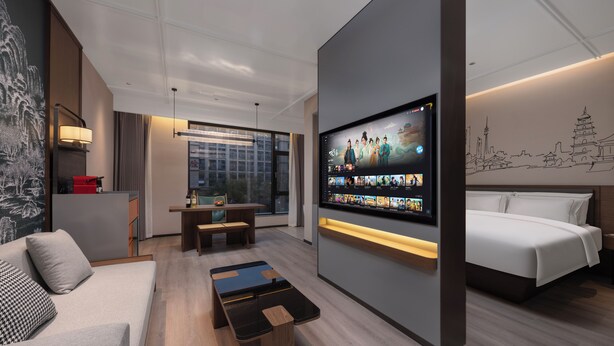 A modern hotel room with a large TV displaying various content, a sofa, a bed, a small table, and a view of the city through large windows. - UrCove by Hyatt Xi'an Bell Tower Huimin Street (Xi'an)