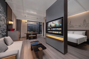 Una habitación de hotel moderna con una televisión grande mostrando diverso contenido, un sofá, una cama, una mesita y vistas de la ciudad a través de amplias ventanas.
