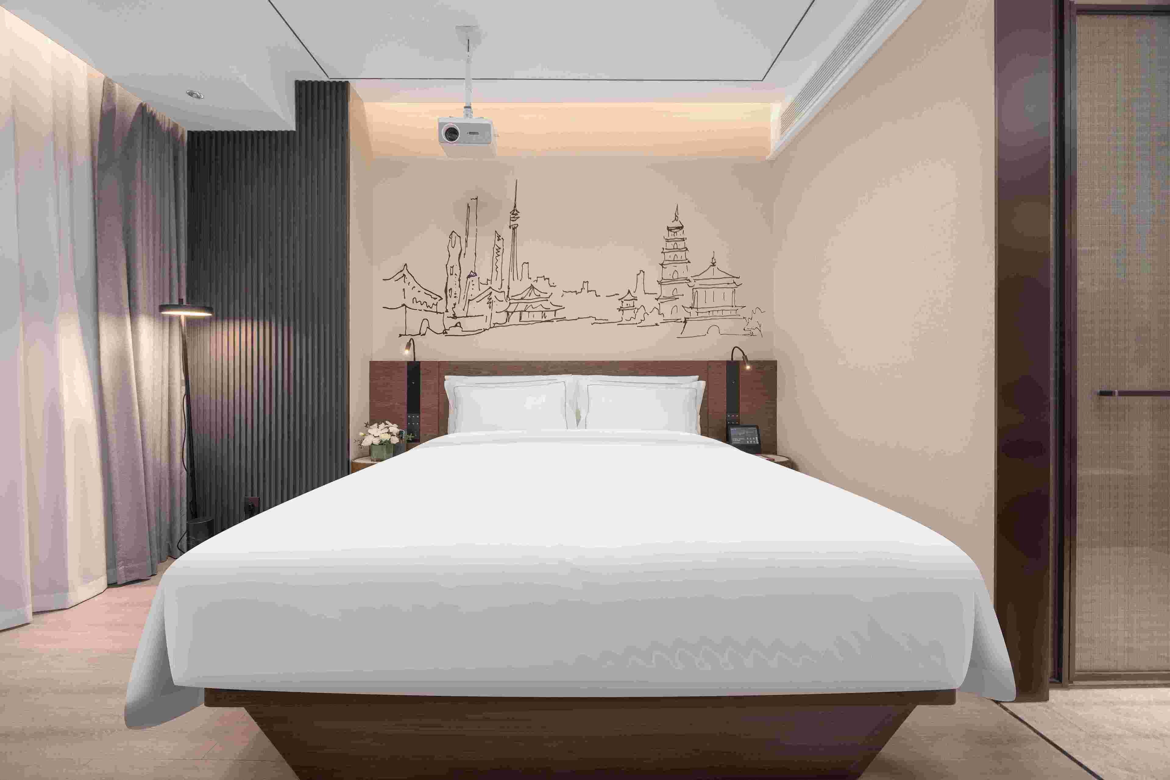 A hotel room with a bed, a wall-mounted projector, and a cityscape mural.